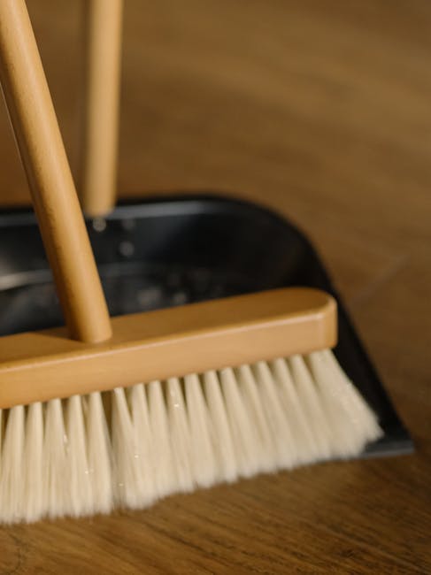 A light blue, irregularly shaped wall hook mounted on a neutral-colored wall in a room, holding a small wooden cleaning brush with stiff bristles and a pink-handled dustpan. The wall surface appears c