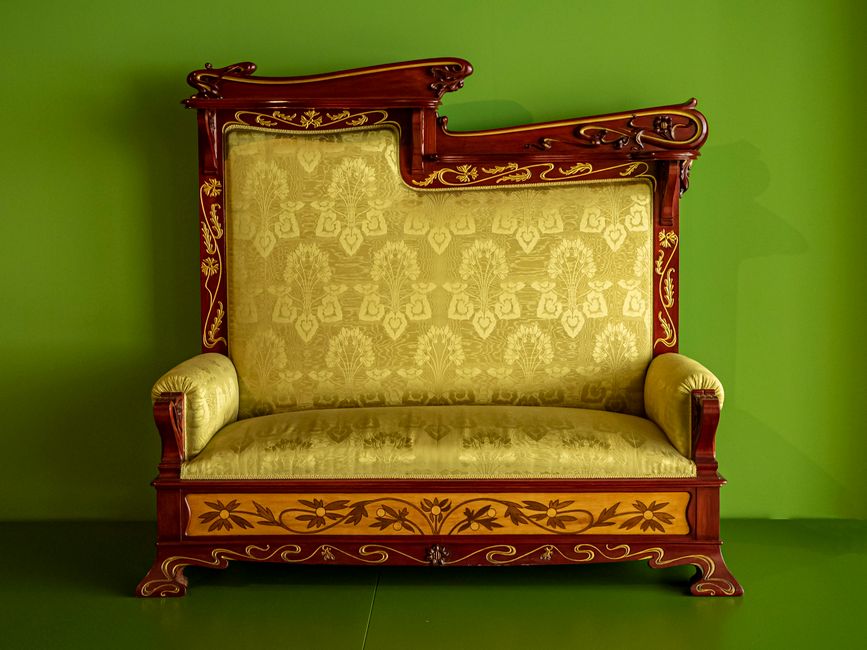 A Victorian-style living room featuring a dark, ornate sofa with carved wooden details and plush upholstery, positioned on a traditional patterned area rug. Behind the sofa, tall windows with sheer wh