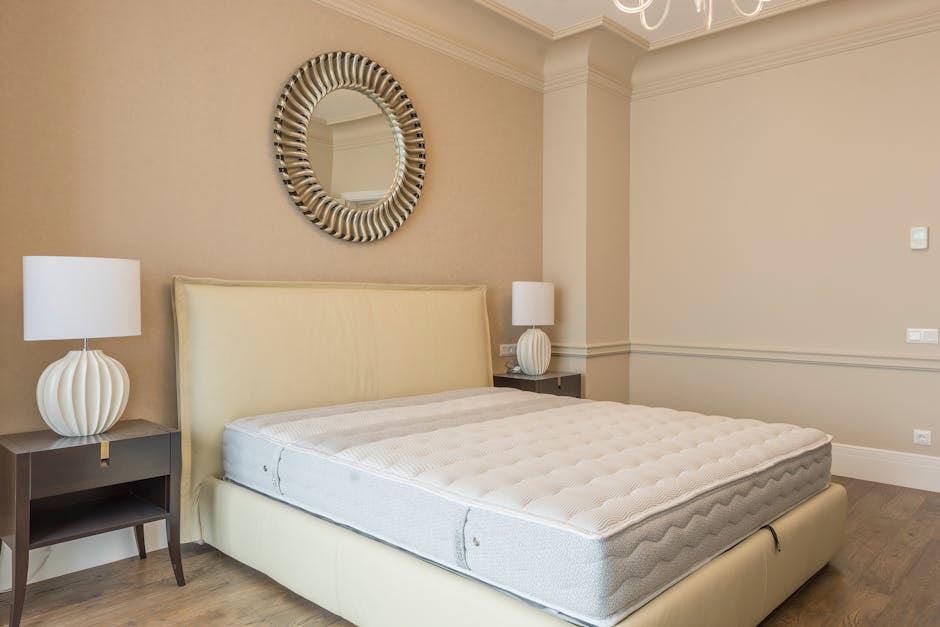 A bedroom featuring a large, white upholstered bed with a new mattress on top, flanked by two dark wooden bedside tables with matching white, rounded ceramic lamps. The wall behind the bed is painted 