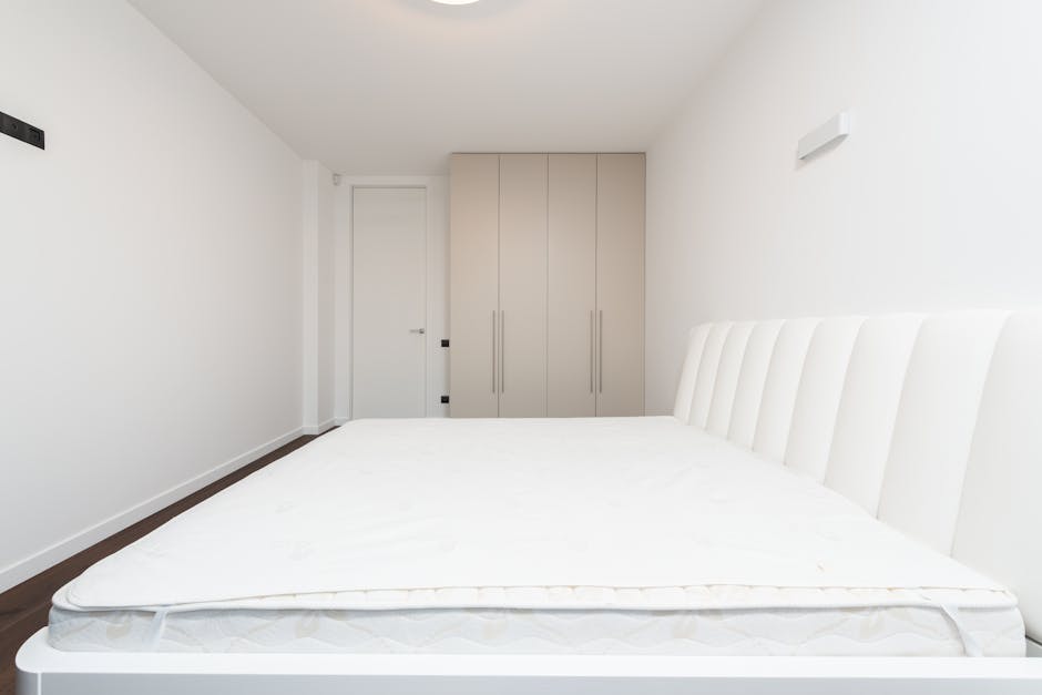 A bedroom featuring a large, white upholstered bed with a new mattress on top, flanked by two dark wooden bedside tables with matching white, rounded ceramic lamps. The wall behind the bed is painted 
